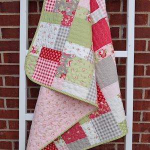 Op de afbeelding: Een patchwork quilt bestaande uit diverse stoffen vierkanten in tinten roze, rood, saliegroen en grijs. Het ontwerp bevat bloemenprints, vichy-ruitjes en effen texturen, afgewerkt met een lichtgroene en witte bies met patroon.