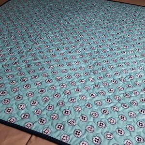 Op de afbeelding: Een turquoise blauwe quilt met een wit en zwart bloemenpatroon. De quilt heeft een donkerblauwe rand.