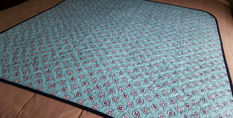 Op de afbeelding: Een turquoise blauwe quilt met een wit en zwart bloemenpatroon. De quilt heeft een donkerblauwe rand.