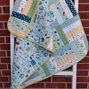 May include: A baby quilt with a patchwork design, featuring various patterns in shades of blue, green, yellow, and white. The quilt has a yellow border and is decorated with cartoon animals and baby-themed illustrations. It is draped over a white ladder.