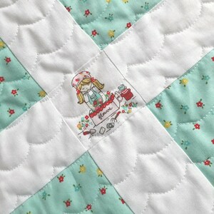 Op de afbeelding: Een wit quilt met een groen bloemenpatroon. Het quilt is gestikt met een gegolfd ontwerp. In het midden van het quilt is een geborduurd beeld van een meisje dat een mand met bloemen vasthoudt. De mand is gelabeld met "Flowers".