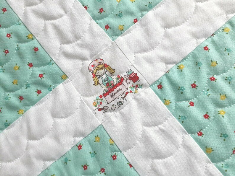 Op de afbeelding: Een wit quilt met een groen bloemenpatroon. Het quilt is gestikt met een gegolfd ontwerp. In het midden van het quilt is een geborduurd beeld van een meisje dat een mand met bloemen vasthoudt. De mand is gelabeld met "Flowers".