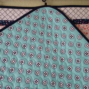 Op de afbeelding: Een turquoise blauwe quilt met een wit en zwart patroon. De quilt is gestikt met een cirkelvormig patroon. De quilt heeft een donkerblauwe bies.