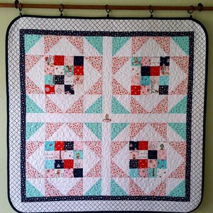 Op de afbeelding: Een wit quilt met een donkerblauwe rand en een patchwork-ontwerp. Het quilt is gemaakt van vierkanten en driehoeken in tinten roze, rood, blauw en wit. Het quilt hangt aan een muur.