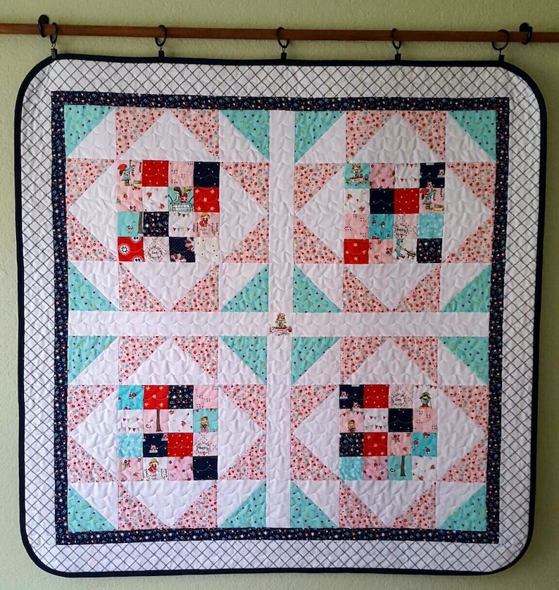 Op de afbeelding: Een wit quilt met een donkerblauwe rand en een patchwork-ontwerp. Het quilt is gemaakt van vierkanten en driehoeken in tinten roze, rood, blauw en wit. Het quilt hangt aan een muur.