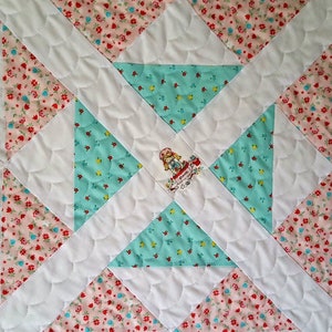 Op de afbeelding: Een wit quilt met een patchwork-ontwerp met roze en blauwe bloemenstof. Het midden van het quilt heeft een witte applicatie van een meisje in een jurk. Het quilt is gestikt met een gegolfd patroon.