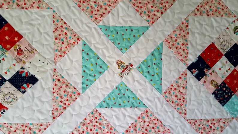 Op de afbeelding: Een wit quilt met een patchwork-ontwerp met roze en blauwe bloemenstof. Het midden van het quilt heeft een witte applicatie van een meisje in een jurk. Het quilt is gestikt met een gegolfd patroon.