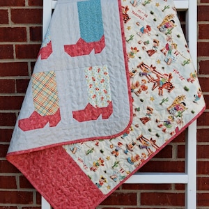 May include: A quilted blanket featuring a pattern of cowboy boots in red, blue, and plaid on a light gray background, contrasted with a cream-colored section printed with cartoon cowboys, cacti, stars, and the text HI-YA PARDNER and YIP-EE.