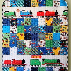May include: A colorful quilt featuring various train designs. The quilt showcases a patchwork design with squares of blue, green, yellow, and white fabric. The top and bottom borders feature train illustrations and the word "SODOR".