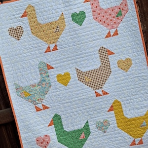 Op de afbeelding: Een witte gewatteerde wandhanger met zes ganzen in diverse stoffen met patronen, waaronder geel, roze, blauw bloemmotief, bruin gingham, groen en effen geel, afgewisseld met kleine stoffen hartjes en een oranje rand.