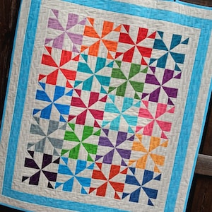 Op de afbeelding: Een rechthoekige quilt met een herhalend windmolenpatroon in diverse kleuren zoals oranje, rood, groenblauw, groen, paars, roze en blauw, op een witte achtergrond met een helder lichtblauwe rand en witte afwerking.