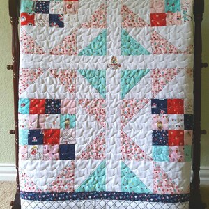 Op de afbeelding: Een wit quilt met een patchwork ontwerp met rode, blauwe en roze bloemenpatronen. Het quilt is gestikt met een golvend patroon en heeft een blauwe en witte rand.