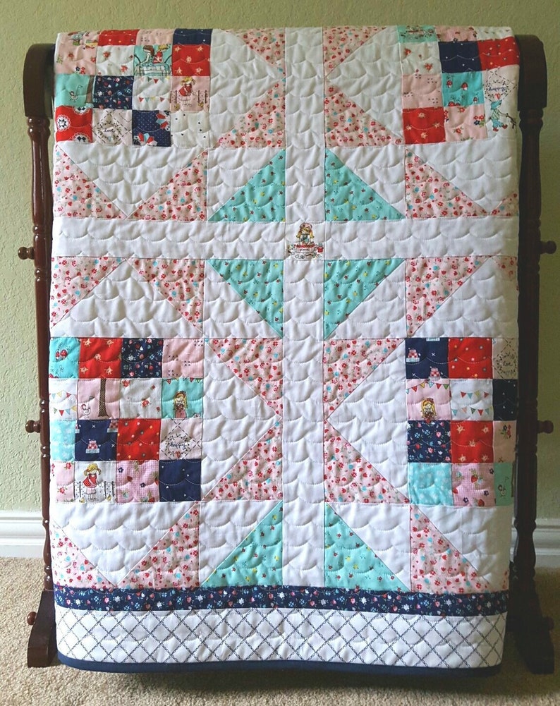 Op de afbeelding: Een wit quilt met een patchwork ontwerp met rode, blauwe en roze bloemenpatronen. Het quilt is gestikt met een golvend patroon en heeft een blauwe en witte rand.