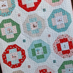 Op de afbeelding: Een doorgestikte deken met een witte achtergrond en meerdere achthoekige blokken in rood, groen, blauw en roze. De blokken bevatten diverse prints met bomen, figuren en geometrische vormen, verbonden door witte stof met een golvend stikselpatroon.