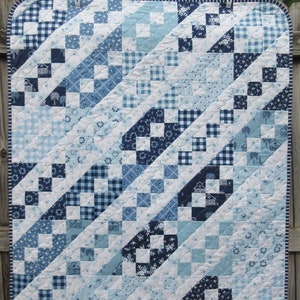 May include: A blue and white quilt with a diagonal pattern. The quilt is made of various fabrics with different patterns, including gingham, floral, and geometric designs. The quilt is hanging on a wooden fence.