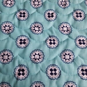 Op de afbeelding: Een turquoise stof met een herhalend patroon van witte en donkerblauwe cirkels. De cirkels hebben een stervormig ontwerp in het midden en een gegolfde rand.