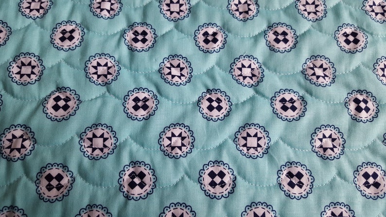 Op de afbeelding: Een turquoise stof met een herhalend patroon van witte en donkerblauwe cirkels. De cirkels hebben een stervormig ontwerp in het midden en een gegolfde rand.
