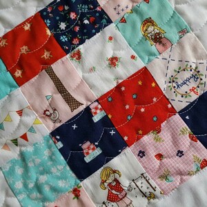 Op de afbeelding: Een patchworkdeken met een verscheidenheid aan kleurrijke stoffen, waaronder rood, blauw, roze en wit. De deken is gemaakt van vierkanten met verschillende patronen, waaronder bloemen, sterren en geometrische vormen. De deken is gestikt met een golvend steekpatroon.