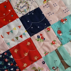 Op de afbeelding: Een patchworkdeken met een verscheidenheid aan kleurrijke stoffen, waaronder rood, roze, blauw en groen. De deken is gemaakt van vierkanten, elk met een ander patroon. De deken is gestikt met wit garen, waardoor een golvend patroon ontstaat.