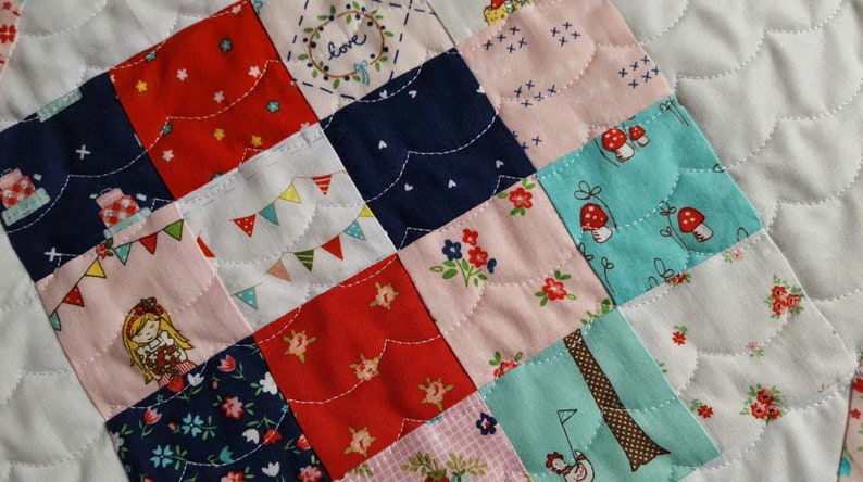 Op de afbeelding: Een patchworkdeken met een verscheidenheid aan kleurrijke stoffen, waaronder rood, roze, blauw en groen. De deken is gemaakt van vierkanten, elk met een ander patroon. De deken is gestikt met wit garen, waardoor een golvend patroon ontstaat.