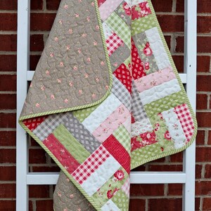 Op de afbeelding: Een doorgestikte deken met een patchworkontwerp van rechthoekige stroken stof in rood, roze, groen en wit. De patronen bevatten bloemenprints, vichy-ruitjes en effen kleuren. De achterkant is van een effen taupekleurige stof met kleine roze roosjes.