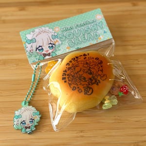 May include: A clear plastic bag contains a yellow squishy bread with a black illustration and text. A matching charm with a cartoon character is attached. The packaging includes the text "Mini Fantôme Squishy Bread."