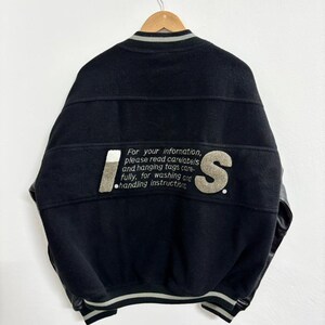 May include: Black varsity jacket with the letters "IS" in a gray and white design on the back. The jacket has a ribbed collar, cuffs, and hem with gray and white stripes. The text "For your information, please read care labels and hanging tags carefully, for washing and handling instructions" is also visible.