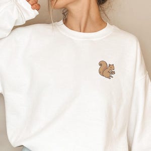 May include: White long-sleeved sweatshirt with a small embroidered squirrel design on the chest. The squirrel is brown and holding a nut. The sweatshirt is a casual style, suitable for everyday wear.