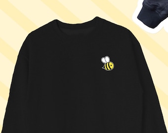 Könnte beinhalten: Schwarzer Pullover mit einem kleinen gelben Bienen-Motiv auf der linken Brust.