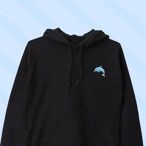 Puede incluir: Sudadera con capucha negra con un pequeño gráfico de delfín azul en la parte delantera. La sudadera tiene un bolsillo canguro y una capucha con cordón.