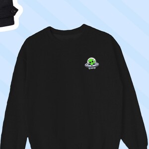 Puede incluir: Sudadera negra de manga larga con un extraterrestre verde en un platillo volante gráfico en el pecho izquierdo.