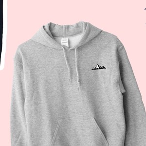 Op de afbeelding: Een lichtgrijze hoodie met een zwart bergmotief op de linkerbovenborst. De hoodie heeft een voorzak, een capuchon met trekkoord en lange mouwen. De achtergrond is lichtroze met decoratieve vormen en andere kledingstukken.