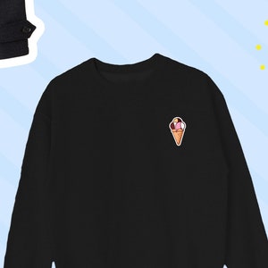 Op de afbeelding: Zwart sweatshirt met ronde hals en een klein geborduurd ijsje op de linkerbovenborst. De ijshoorntje heeft bolletjes roze, witte en bruine ijs in een wafel.