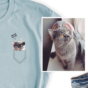 May include: Light blue t-shirt featuring a graphic of a cat wearing sunglasses in a pocket design. The cat has the text "HERE FOR THE FOOD" above its head. A separate image shows a gray cat with orange eyes.