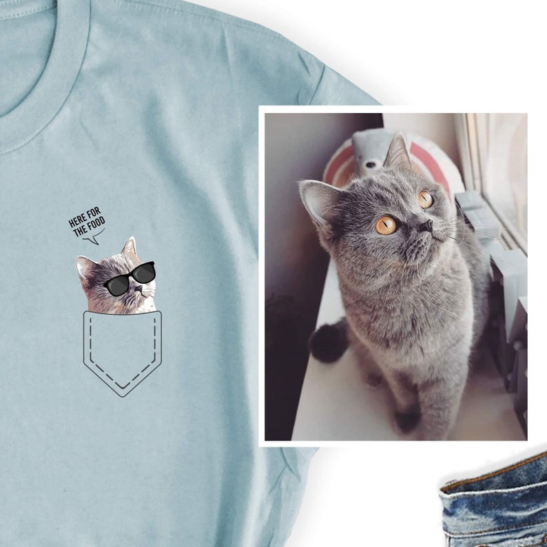 May include: Light blue t-shirt featuring a graphic of a cat wearing sunglasses in a pocket design. The cat has the text "HERE FOR THE FOOD" above its head. A separate image shows a gray cat with orange eyes.
