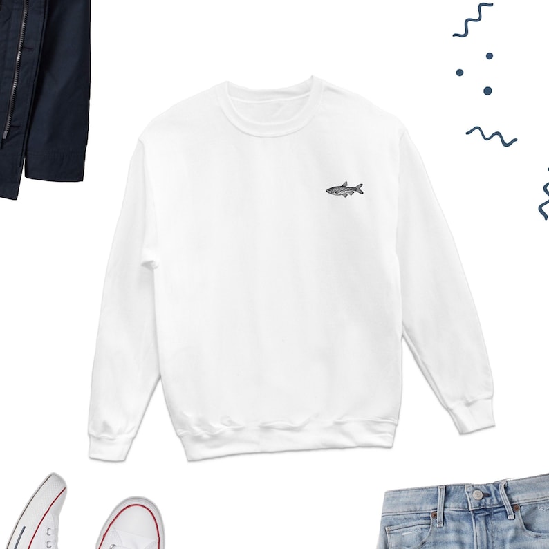 K&ouml;nnte beinhalten: Wei&szlig;er Langarm-Pullover mit einem kleinen gestickten Fischmotiv auf der Vorderseite.