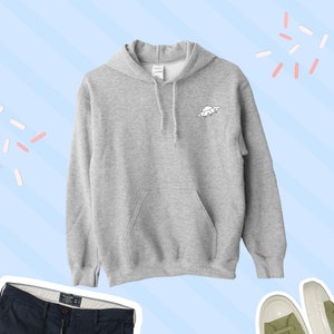 K&ouml;nnte beinhalten: Ein grauer Kapuzenpullover mit einem kleinen schwarzen und wei&szlig;en UFO-Motiv auf der Vorderseite. Der Pullover hat eine K&auml;ngurutasche und eine Kapuze mit Kordelzug.