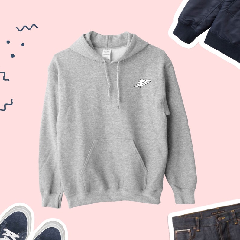 K&ouml;nnte beinhalten: Ein grauer Kapuzenpullover mit einem kleinen wei&szlig;en UFO-Motiv auf der Vorderseite. Der Pullover hat eine K&auml;ngurutasche und eine Kapuze mit Kordelzug.