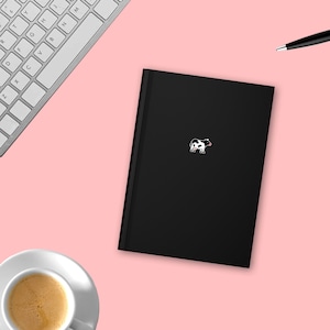 Puede incluir: Un cuaderno negro con una pequeña pegatina de vaca blanca en la parte delantera. El cuaderno está sobre una superficie rosa.