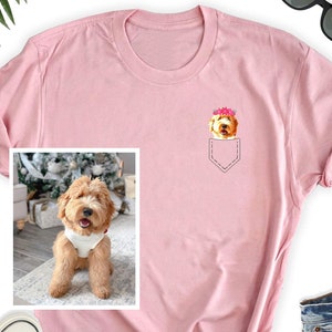May include: A light pink t-shirt featuring a pocket design with a cartoon dog wearing a flower crown. The dog is light brown with a white chest. A framed photo of the dog is on the left side of the shirt.