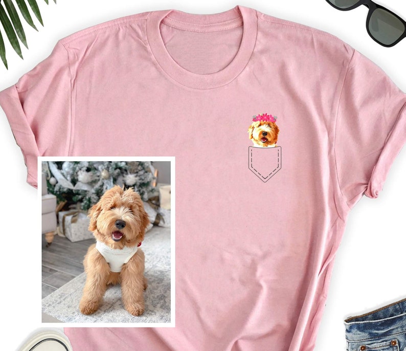 May include: A light pink t-shirt featuring a pocket design with a cartoon dog wearing a flower crown. The dog is light brown with a white chest. A framed photo of the dog is on the left side of the shirt.