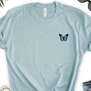 May include: A light blue t-shirt with a small blue butterfly graphic on the left chest.