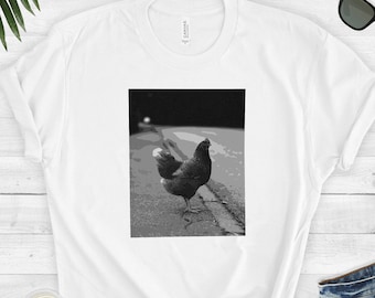Pode incluir: Uma camiseta gráfica em preto e branco com uma galinha em pé sobre uma superfície texturizada com um fundo escuro.