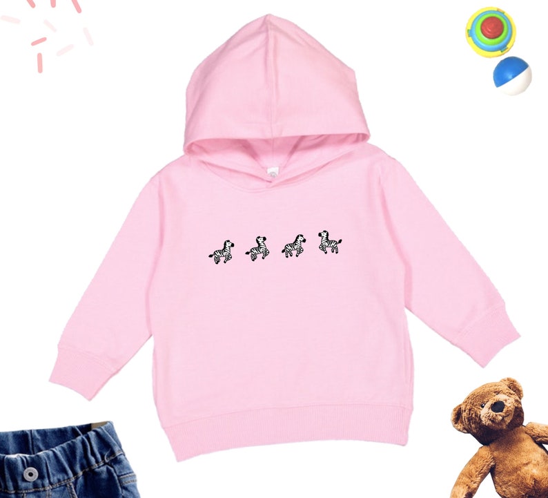 May include: A pink hooded sweatshirt with a graphic of four zebras on the front. The sweatshirt has long sleeves and a kangaroo pocket.