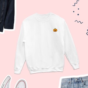 Puede incluir: Sudadera blanca de manga larga con una peque&ntilde;a calabaza bordada en la parte delantera. La calabaza es naranja con un tallo verde y una cara negra.