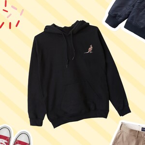 May include: Black hoodie featuring a kangaroo emblem on the chest. The kangaroo is illustrated in brown and beige tones. The sweatshirt includes a front pocket and a drawstring hood. The background has a yellow and white striped design.