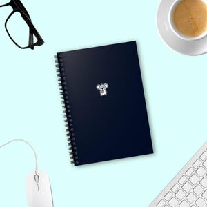 Może przedstawiać: Czarny notes w spirali z małą naklejką z koalą z przodu. Notes leży na jasnoniebieskiej powierzchni z białym kubkiem do kawy i białą klawiaturą.