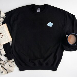 May include: A black sweatshirt with a small blue cloud with a smiling face embroidered on the left chest. The sweatshirt is laying flat on a white surface with a book, glasses, and a scarf beside it.