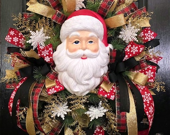 May include: A decorative wreath featuring a central molded Santa Claus face with a white beard and red hat. The wreath is adorned with green pine branches, gold glittered sprigs, white snowflake ornaments, and ribbons in red, black, gold, and red plaid patterns.