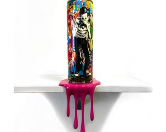 May include: A cylindrical spray paint can featuring a black and white graphic of a person holding a camera, set against a vibrant, multi-colored splattered background. A thick, glossy pink substance drips from the base of the can.